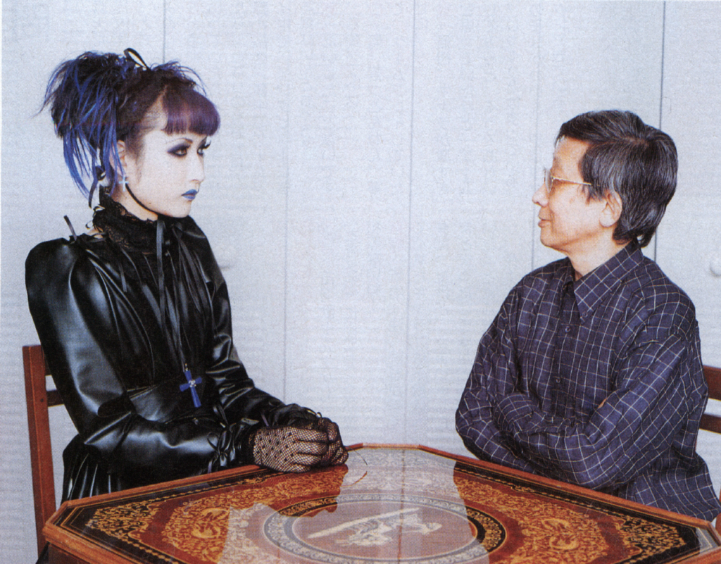 Mana from Malice Mizer and Koichi Sugiyama looking at each other, sitting at a table
